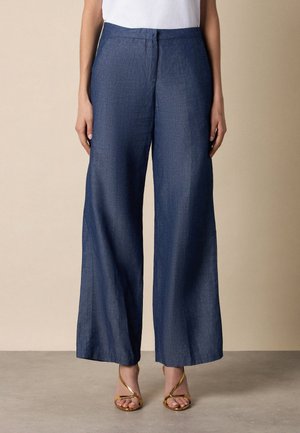 Kobieta ubrana w ciemnoniebieskie, szerokie spodnie z wysokim stanem, w połączeniu ze złotymi sandałami na obcasie z paskami, stojąca na beżowym tle.