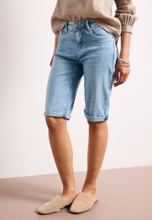 Person iført lyseblå, opkørte denim Bermuda-shorts, beige langærmet skjorte, sølv armbånd og beige slip-on sko.