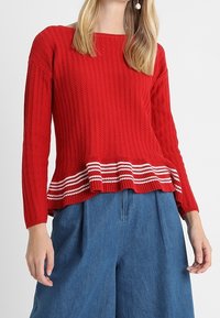 Red textured knit sweater with long sleeves, featuring a ruffled hem accented by white and red stripes, paired with blue wide-leg trousers.