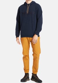 Marineblauwe hoodie met reliëf Timberland-logo, kangoeroezak, gecombineerd met mosterdgele broek met elastische cuffs en donkere sneakers.