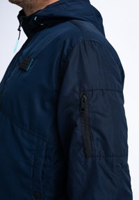 Námořnická modrá bunda s kapucí, vyrobená z hladkého, voděodolného materiálu. Obsahuje kapsu na zip na rukávu a logo na hrudi.