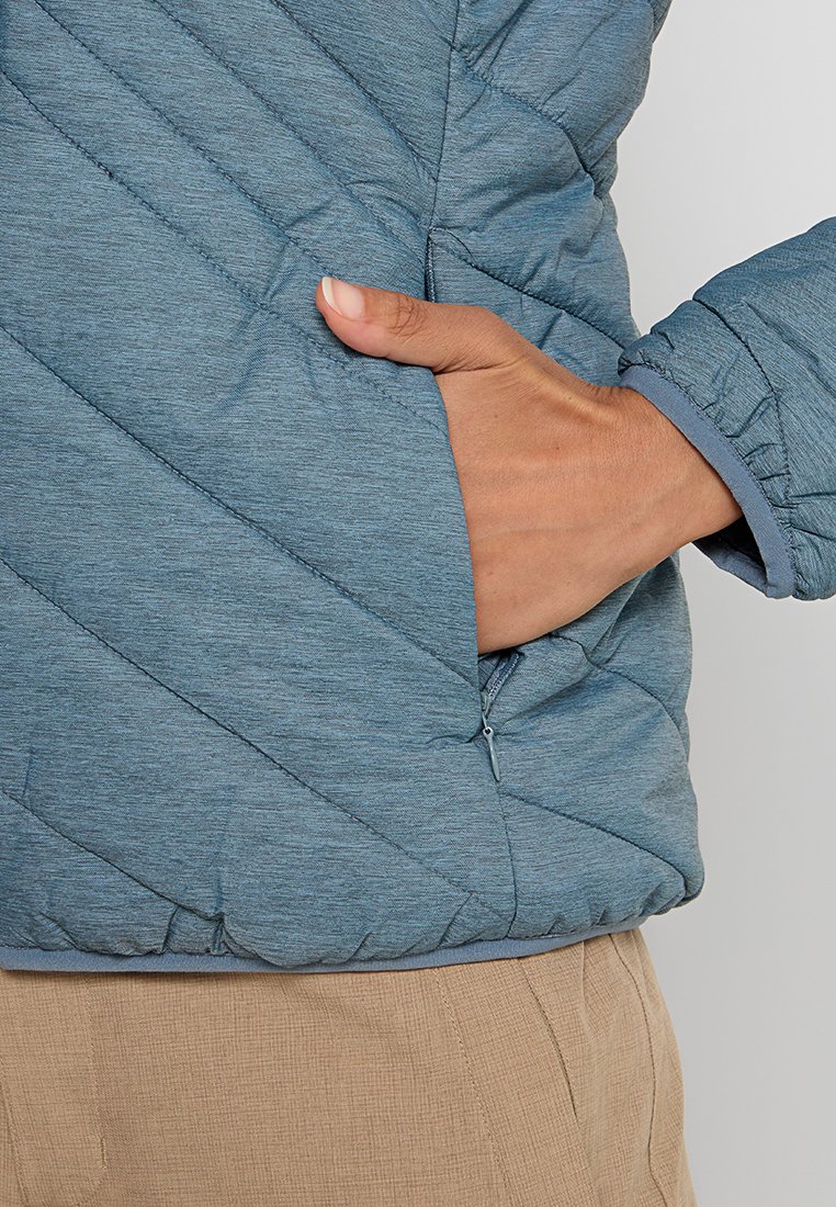 Himmelblau gesteppte Jacke mit diagonaler Nahtmuster, ausgestattet mit einer Seitentasche und gerippten Bündchen. Das Material wirkt weich und strukturiert.