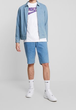 Veste en denim bleu clair avec fermeture éclair, t-shirt blanc avec un logo Nike violet, shorts bleu clair et baskets blanches avec des chaussettes mi-mollet.