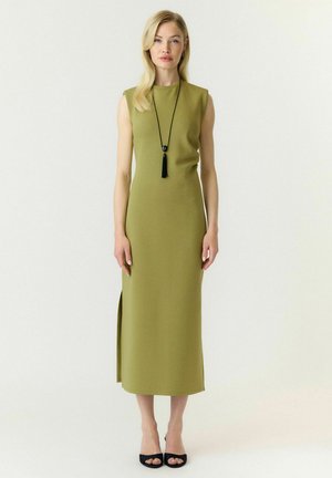 Femme portant une robe midi olive sans manches, un collier noir à pompons et des talons noirs ouverts, debout devant un fond uni.