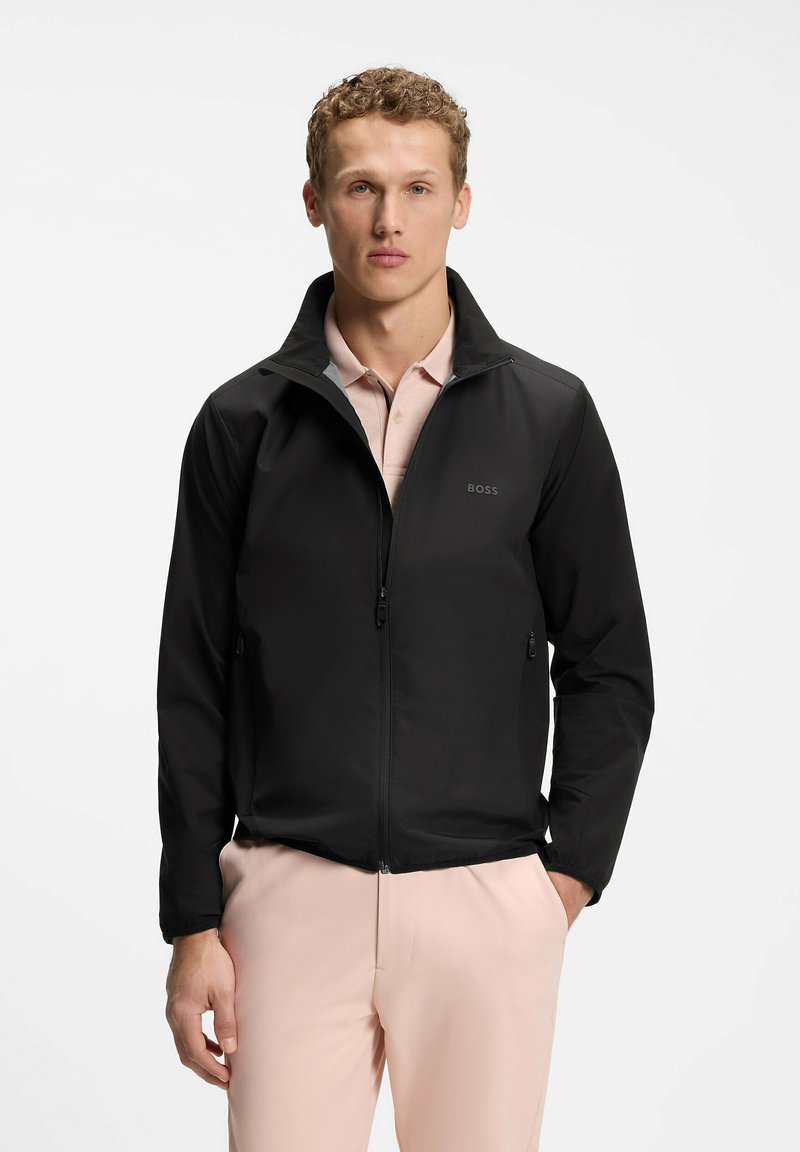 Jeune homme portant une veste BOSS noire zippée sur un polo rose clair et un pantalon rose assorti, debout les mains dans les poches.