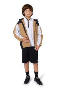 Giacca a vento zipata beige e nera con dettagli bianchi, indossata sopra un pullover bianco. Pantaloni corti neri, calze nere e scarpe da ginnastica nere completano l'outfit.