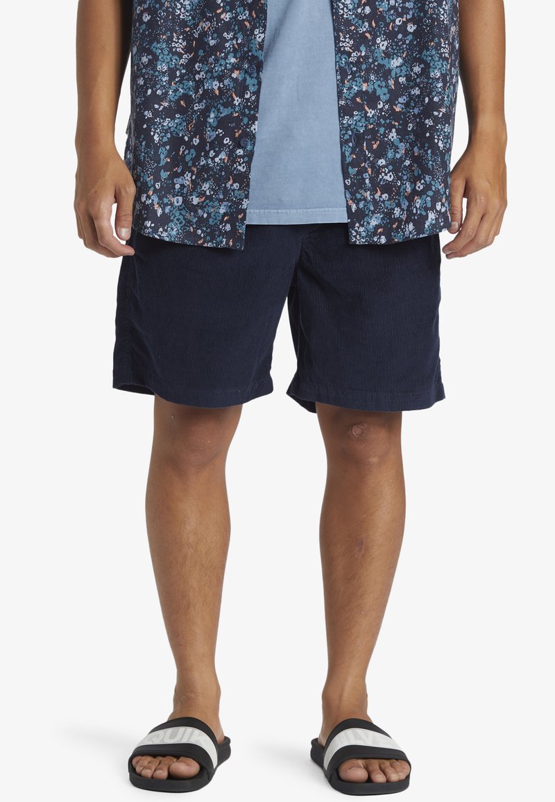 Kortermet blomsterskjorte i mørk blå og tealfarge, sammen med marineblå kordfløyelsshorts. Svart sandaler med hvite striper på føttene.