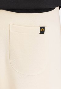 Cremefarvede sweatpants har en stor baglomme med afrundede kanter, fremhævet af et sort mærke med en gul logo detail.