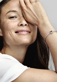 Pulsera de corazón plateada en la muñeca de una mujer, con un diseño elegante y texturizado. La mujer tiene el cabello largo y pecas, y lleva una camisa blanca.