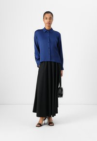 Chemise boutonnée bleu à manches longues, coupe ample, associée à une jupe maxi plissée noire. Un sac à main noir et des sandales à talons complètent le look.