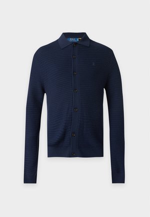 Jeune homme debout, les mains dans les poches, portant un cardigan bleu marine boutonné, un pantalon marron à carreaux et des baskets blanches, sur un fond clair uni.