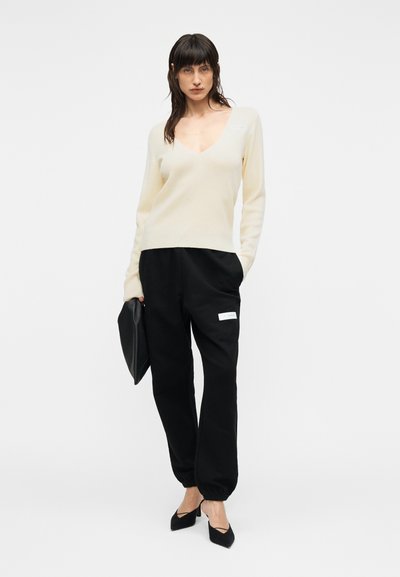 Κρεμ πουλόβερ με V λαιμόκοψη σε συνδυασμό με μαύρα παντελόνια jogger με πλαϊνή τσέπη. Το μοντέλο κρατά μαύρο clutch και φορά μαύρες μυτερές γόβες.