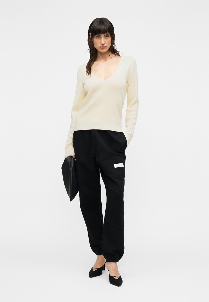 Pull crème à col en V associé à un pantalon de jogging noir avec une poche latérale. Le mannequin tient une pochette noire et porte des escarpins noirs à bouts pointus.