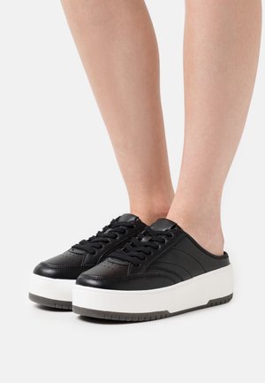 Personne portant des baskets noires sans lacets avec des semelles blanches épaisses, montrant les jambes inférieures sur un fond clair uni.