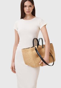 Borsa a spalla in paglia intrecciata con superficie testurizzata, dotata di manici in pelle nera e apertura superiore, tenuta da una persona in un vestito chiaro.