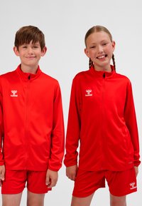 Due bambini sorridenti con giacche sportive rosse abbinate e pantaloncini, in piedi fianco a fianco contro uno sfondo uniforme.