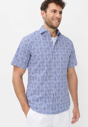 Mann lächelt, trägt ein blau-weiß vertikal gestreiftes Hemd mit kurzen Ärmeln und weiße Hose, Hände in den Taschen, vor einem schlichten Hintergrund.