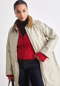 Beige coat with a brown corduroy collar, worn over a red ribbed cardigan, paired with dark jeans. Features a zip closure and pockets.