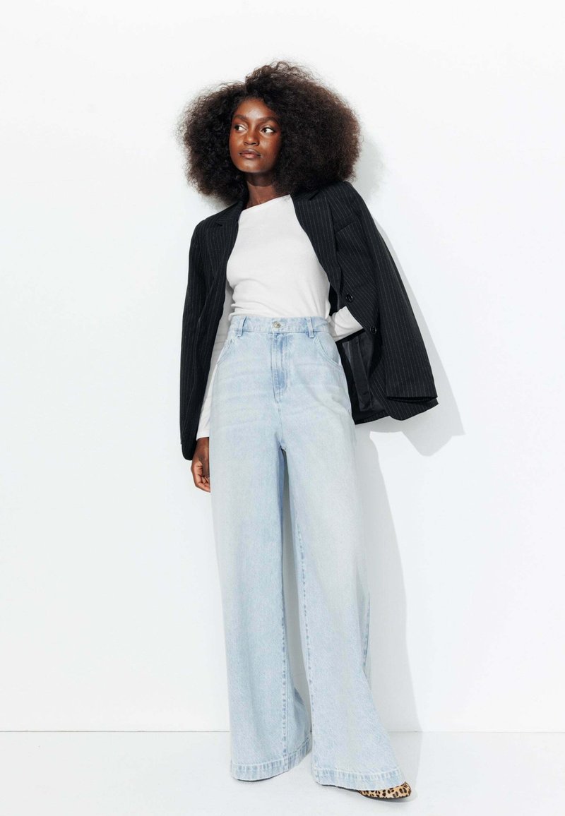 Blazer noir à rayures, chemise blanche ajustée, jeans taille haute bleu clair à jambes larges, et talons léopard. Texture lisse et coupe moderne.