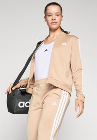 Mlada ženska v bež Adidas trenirki in belem topu, ki nosi črno Adidas torbo preko ramena, stoji samozavestno.