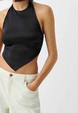 Woman wearing a black satin halter top with pointed hem paired with off-white high-waisted jeans, hand in pocket against plain background.