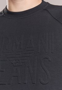 Black sweatshirt with a round neckline featuring embossed "Armani Jeans" lettering and a winged logo. Made of a smooth, textured fabric.