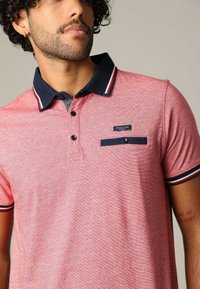 Rood gestreept polo shirt met een marineblauwe kraag, een drieknopen-sleuf en een zak met een blauwe accent. Merkenlabel zichtbaar op de borst.