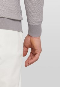 Suéter gris claro con textura y puño acanalado. Una mano se encuentra cerca de una pierna de pantalón blanca, mostrando la longitud y el ajuste de la manga.