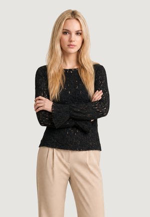Giovane donna con lunghi capelli biondi che indossa un top nero in pizzo a maniche lunghe e pantaloni beige, in piedi con le braccia incrociate su uno sfondo semplice.