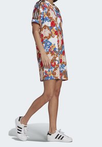 Korte jurk met bloemenpatroon in het wit, met blauwe en rode accenten, voorzien van zwarte mouwen met drie strepen en een ontspannen pasvorm. Gecombineerd met witte sneakers.
