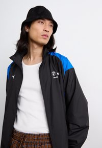 Crni windbreaker s plavim akcentima, s visokim ovratnikom i Puma logom, nosi se preko bijele rebraste majice i smešnih kariranih hlača.