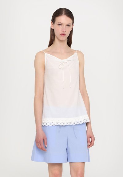 White camisole with a v-neckline and front tie detail, featuring a lace trim at the hem. Paired with light blue pleated shorts.