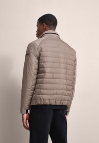 Beige gepolsterte Jacke mit Stehkragen, versehen mit horizontalen Nähten und einem schwarzen Akzent am Ärmel. Glattes, leichtes Material.