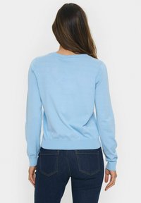 Világoskék kötött pulóver hosszú ujjal és kerek nyakkivágással, szabott alsó szegéllyel. Sötét denim farmernadrággal párosítva.