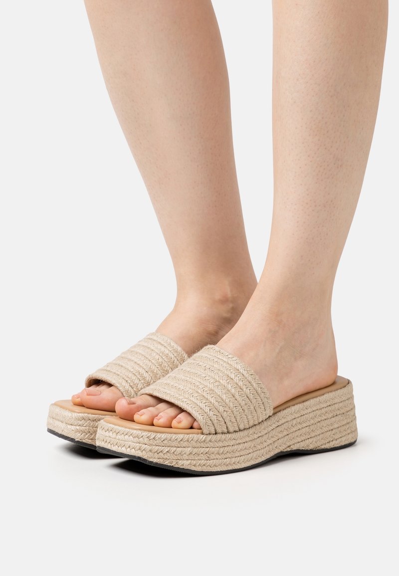 Pieds portant des sandales slides beiges à plateforme avec de larges brides tissées et une texture tressée sur la semelle, présentés sur un fond uni.