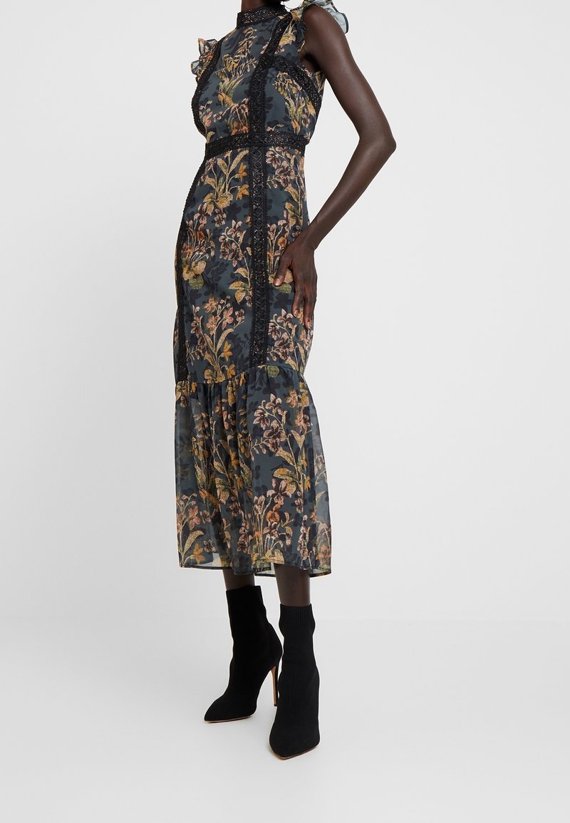 Floral Midi-Kleid aus durchscheinendem schwarzem Stoff mit bunten Blumenmustern, Rüschen an den Schultern und Spitzenverzierungen an der Taille. Kombiniert mit schwarzen Knöchelstiefeln.