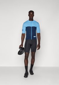 Maillot de cyclisme en bleu clair et bleu marine avec une fermeture éclair à l'avant. Porté avec un short noir et tenant un casque noir. Texture de tissu lisse.