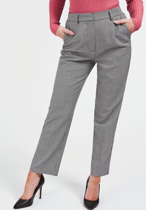 Mujer con pantalones de pierna recta a cuadros gris y blanco, tacones altos negros puntiagudos y una blusa rosa de manga larga acanalada, con las manos en los bolsillos.