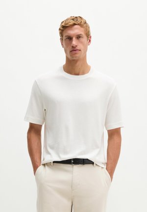 Blonde man draagt een effen wit T-shirt met korte mouwen, beige broek en een zwarte riem, staand met zijn handen in de zakken.