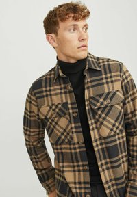 Brązowa i czarna koszula flanelowa w kratę z dwoma kieszeniami na piersi, zapinana na guziki, o miękkiej teksturze, noszona na czarnym prążkowanym golfie.