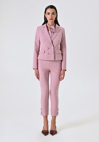 Roze op maat gemaakte blazer met dubbele knopen en bijpassende aansluitende broek. Sjaaldetail bij de hals, kortere lengte en knoopaccenten bij de mouwen.