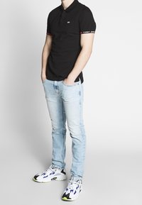 Svart pikétröja med röda detaljer, ljusblå jeans och vita sneakers med blåa och gröna inslag. Enkelt mönster, åtsittande silhuett.