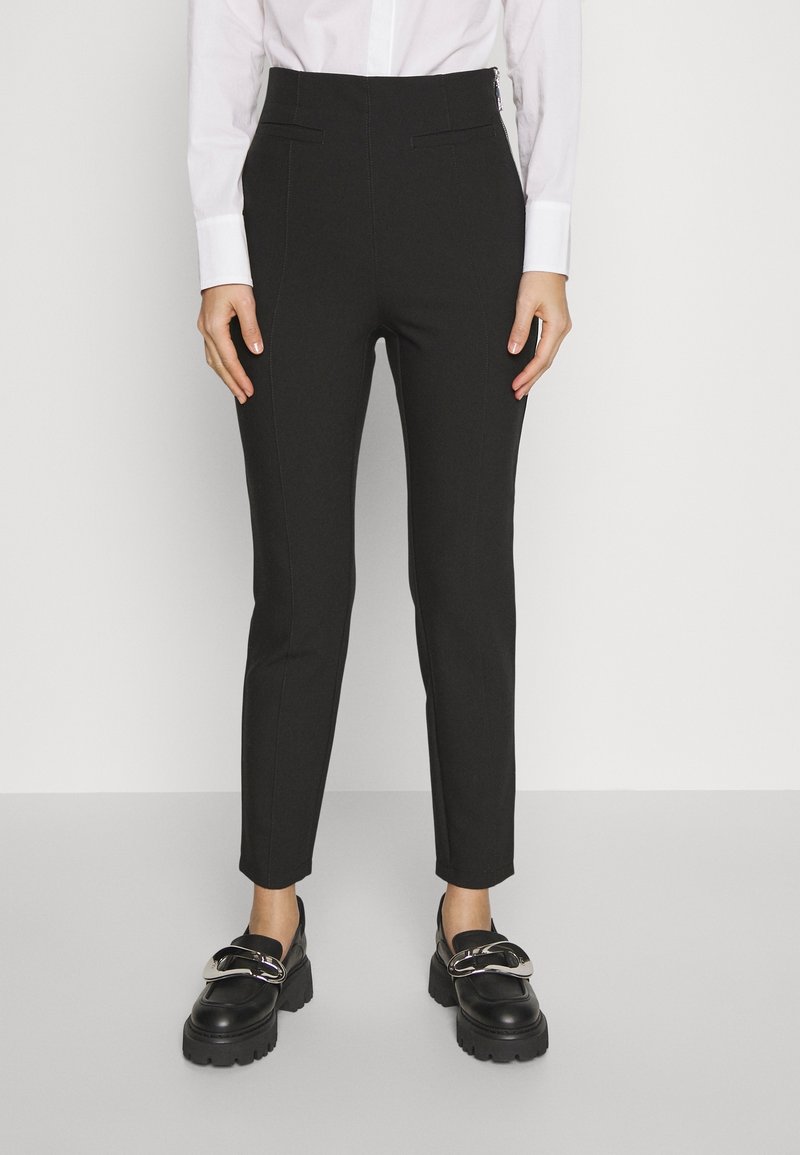 Pantalon noir sur mesure avec taille haute, coupe slim, devant plat avec poches et texture lisse, associé à des mocassins noirs épais.