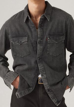 Person trägt ein ausgewaschenes schwarzes Jeanshemd mit Knöpfen und Brusttaschen, die Hände in den Taschen, ein weißes Unterhemd und einen schwarzen Gürtel mit goldener Schnalle.