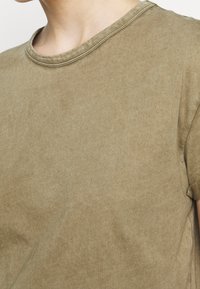 Oliwkowa, krótko rękawowa koszulka z okrągłym dekoltem, wykonana z miękkiego, teksturowanego materiału, z delikatnymi różnicami w kolorze.