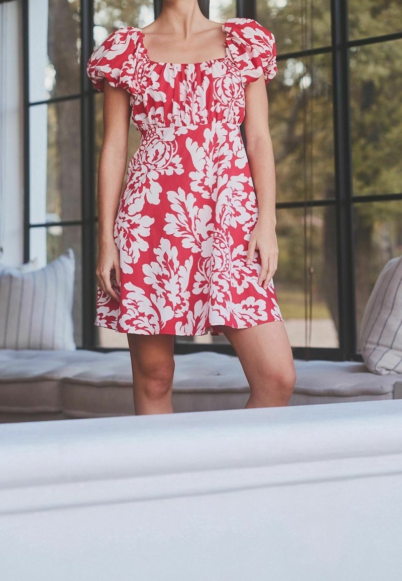 Femme debout à l'intérieur portant une robe rouge à motif floral blanc et manches courtes bouffantes, avec de grandes fenêtres et un canapé en arrière-plan.