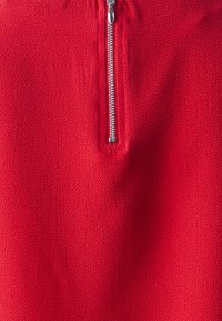 Tessuto rosso con una finitura testurizzata, caratterizzato da una chiusura centrale in metallo argentato, progettato per un facile indossamento e stile.