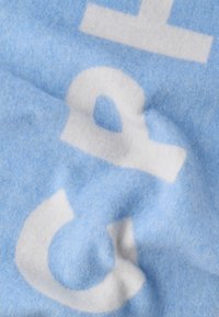 Jasnoniebieski koc o miękkiej, puszystej fakturze z białymi abstrakcyjnymi kształtami i literami. Materiał wydaje się gruby i przytulny.