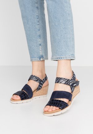 Pieds portant des sandales compensées bleu marine avec des brides à motifs aux chevilles, associées à un jean léger bleu clair coupé court.