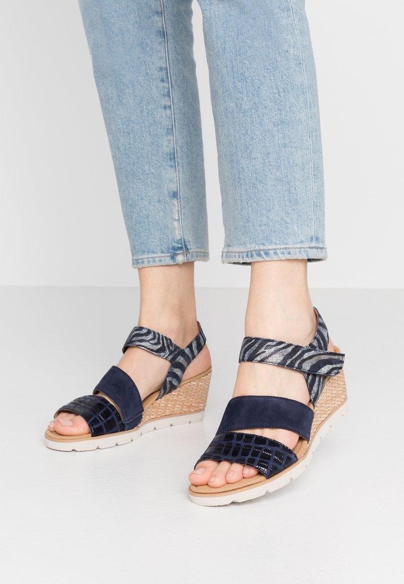 Pieds portant des sandales compensées bleu marine avec des brides à motifs aux chevilles, associées à un jean léger bleu clair coupé court.